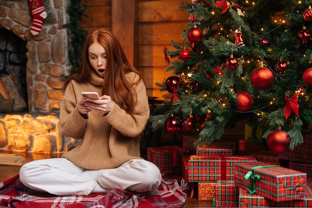 Portret van een verraste roodharige jonge vrouw die een wintertrui draagt en goed nieuws krijgt op de mobiele telefoon