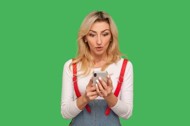 Portret van een verraste volwassen mooie vrouw in een stijlvolle overall die een bericht op een smartphone leest en verbazing uitdrukt, met behulp van een mobiel apparaat voor communicatie. studio-opname geïsoleerd op groene achtergrond