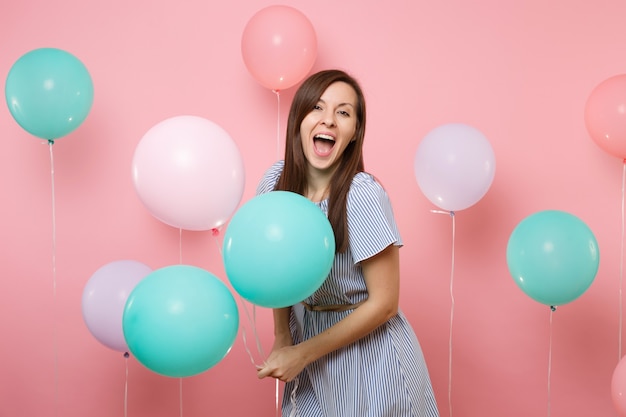 Portret van een verraste vrolijke vrouw met geopende mond in een blauwe jurk met kleurrijke luchtballonnen op een felgekleurde roze achtergrond. Verjaardagsfeestje, mensen oprechte emoties concept.