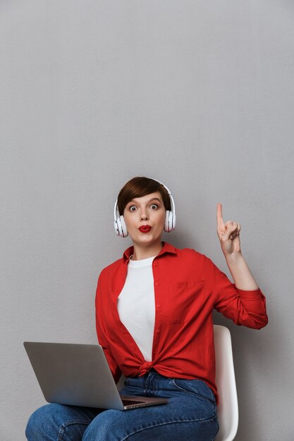 Portret van een verraste vrouw in een koptelefoon die lacht terwijl ze op een stoel zit met een laptopcomputer die over een grijze muur is geïsoleerd