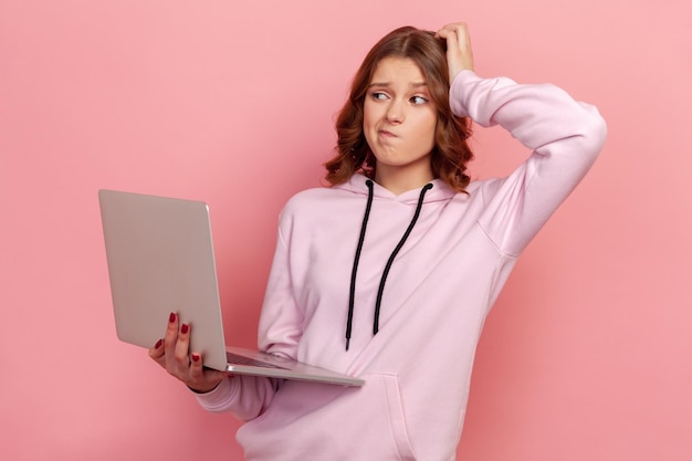 Portret van een verward tienermeisje met krullend haar in hoodie die laptop vasthoudt, hoofd krabt en nadenkt over een nieuw idee. Indoor studio-opname geïsoleerd op roze background