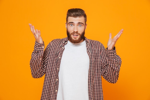 Portret van een verwarde jonge man die vrijetijdskleding draagt die zich geïsoleerd over gele muur bevindt, schouders ophaalt