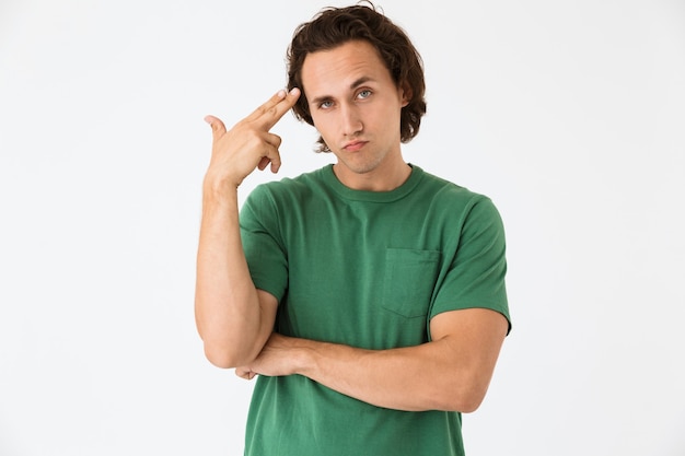 Foto portret van een verwarde jonge man met een basic t-shirt met geweervingers in zijn tempel geïsoleerd over een witte muur