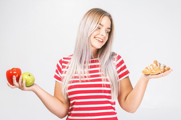 Portret van een verwarde mooie jonge blonde vrouw die tussen gezond en ongezond voedsel kiest. Geïsoleerd op witte achtergrond.