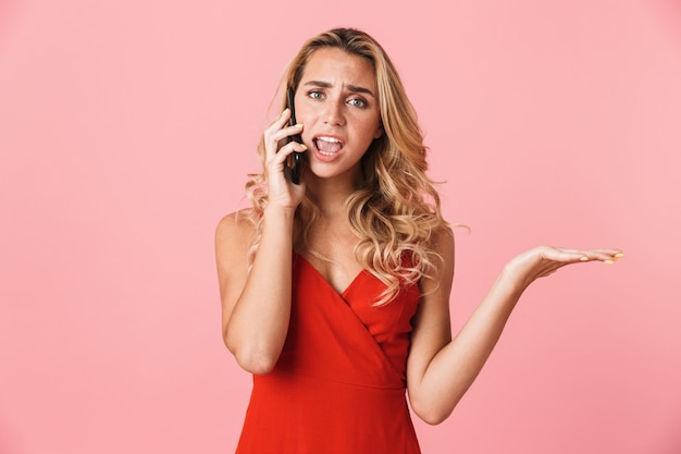 Portret van een verwarde, ontstemde, gestresste jonge blonde vrouw die zich geïsoleerd over een roze muur aan het praten is via de mobiele telefoon