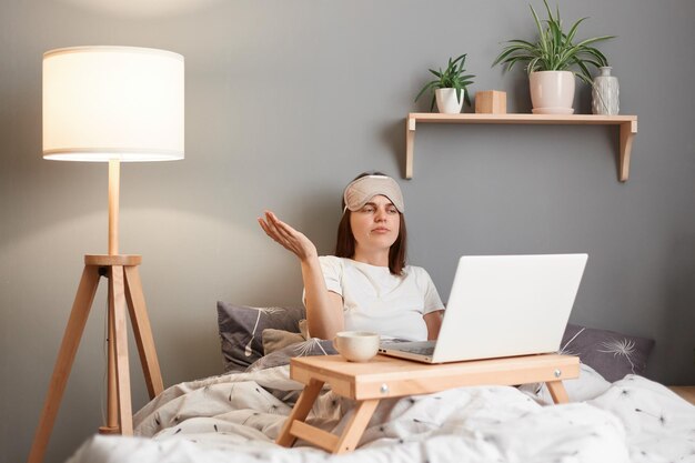 Portret van een verwarde, onzekere vrouw met een slaapmasker die vanuit huis op een laptop werkt terwijl ze op bed ligt en een videogesprek heeft met een partner die online vergadert, weet niet wat ze moet doen en spreidt haar hand opzij