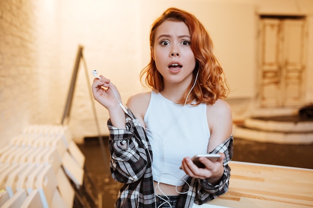 Portret van een verwarde roodharige jongedame gekleed in een shirt in een kooiprint en een wit t-shirt terwijl ze naar muziek luistert en aan het chatten is.