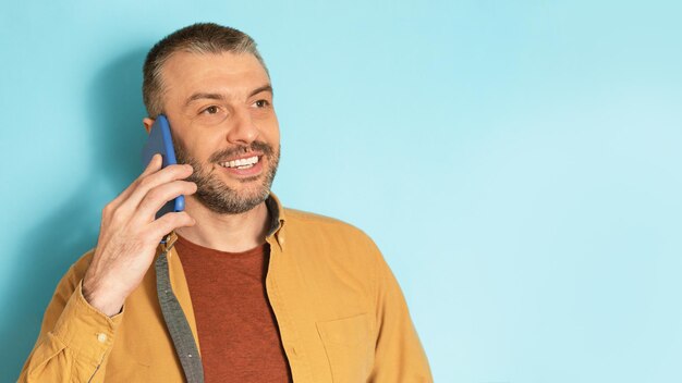 Portret van een volwassen man die op zijn smartphone praat en naar kopieerruimte kijkt die over een blauw studio-achtergrondpanorama staat