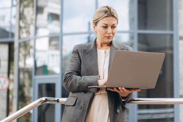 Portret van een volwassen zakenvrouw die lacht met emoties en een laptop vasthoudt die tijdens een videogesprek praat en werk doet op de achtergrond van moderne stedelijke en kantoorgebouwen