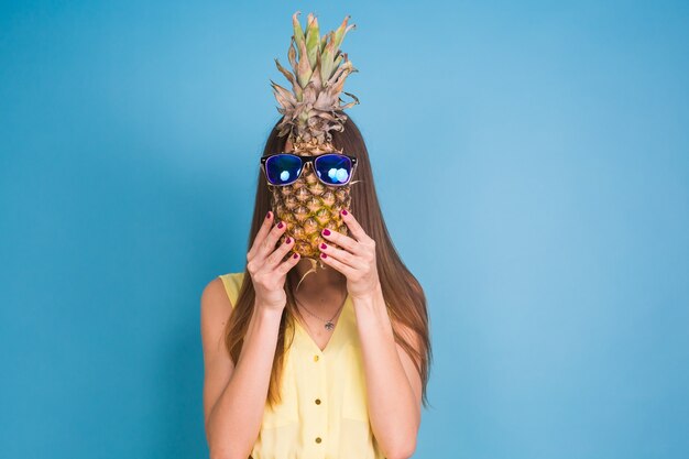 Portret van een vrij grappig zomermeisje die ananas in zonnebril houden die over blauwe achtergrond wordt geïsoleerd.