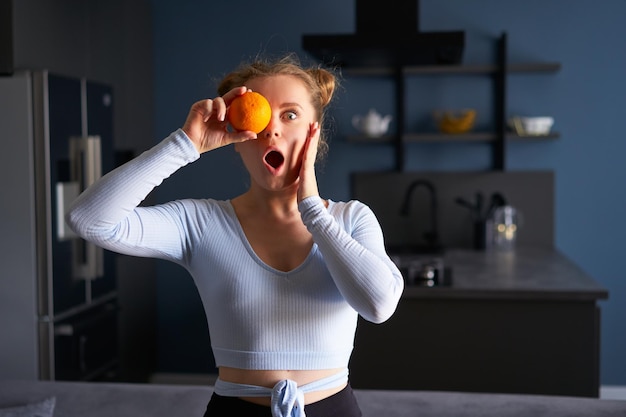 Portret van een vrij Kaukasisch meisje dat een grappig gezicht maakt verrast met verse, sappige sinaasappel voor haar gezicht in de moderne keuken Jonge grimassende vrouw die één oog bedekt met een oranje fruit thuis