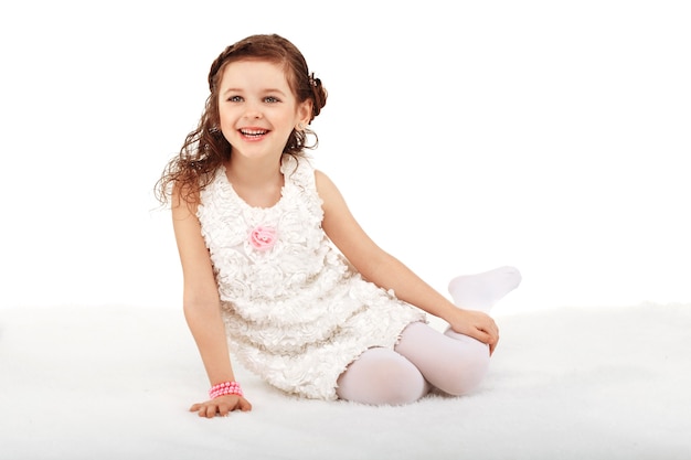 Portret van een vrij klein meisje van de leuke manier, zittend op een zachte deken op de vloer en plezier tegen wit