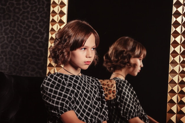 Portret van een vrij schattig tienermeisje in een elegante jurk voor de spiegel in een donker, stijlvol interieur van de woonkamer. Kinderen emoties en poseren. Concept van stijl, mode en schoonheid. Ruimte voor site kopiëren