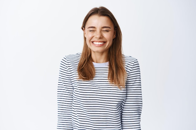 Portret van een vrolijk blond meisje in vrijetijdskleding, plezier maken, zich gelukkig voelen, ogen sluiten en glimlachen met witte tanden, staande op wit