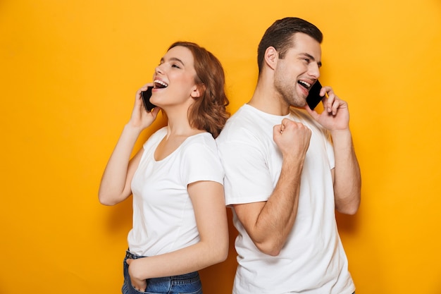 Portret van een vrolijk jong stel dat geïsoleerd over een gele muur staat en mobiele telefoons gebruikt