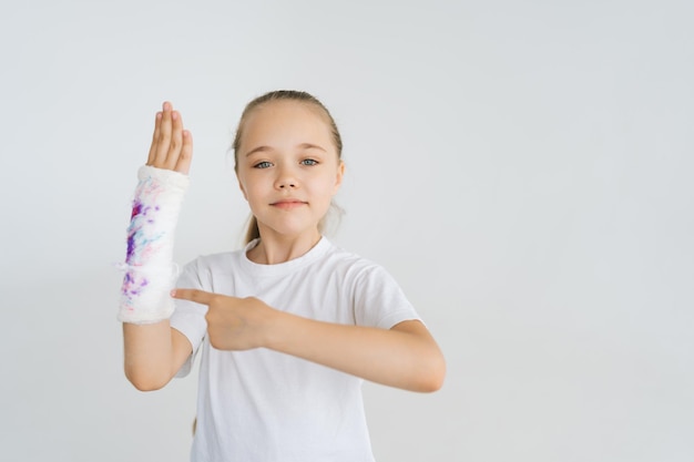 Portret van een vrolijk klein meisje dat met wijsvinger naar gebroken hand laat zien, gewikkeld in wit gipsverband met kleurrijke trekking die naar de camera kijkt