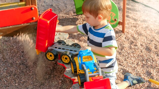 Portret van een vrolijk lachend jongetje dat zand in speelgoedvrachtwagen met aanhanger giet. Kinderen spelen en spelen op de speelplaats in het park