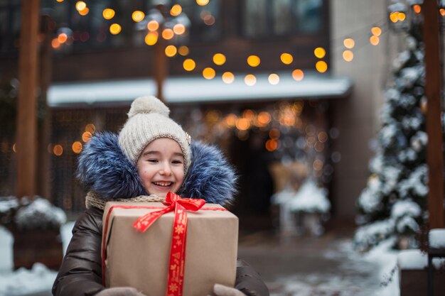 Portret van een vrolijk meisje met een geschenkdoos voor Kerstmis op een stadsstraat in de winter met sneeuw op een feestelijke markt met versieringen en kerstverlichting. Warme kleding, gebreide muts, sjaal en bont. Nieuwjaar