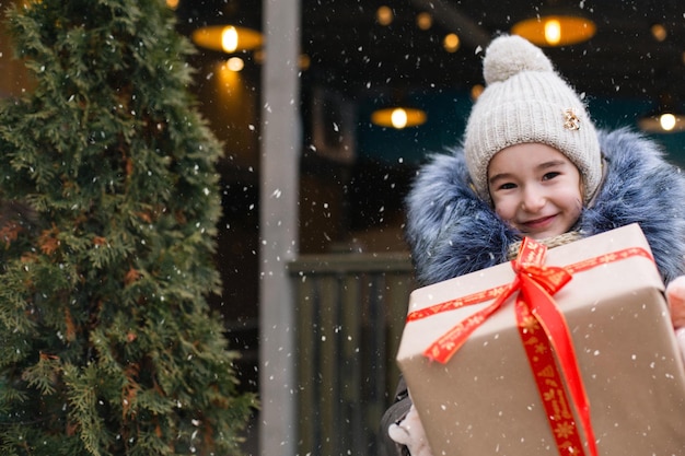 Portret van een vrolijk meisje met een geschenkdoos voor Kerstmis op een stadsstraat in de winter met sneeuw op een feestelijke markt met versieringen en verlichting. Warme kleding, gebreide muts, sjaal en bont. Ruimte kopiëren