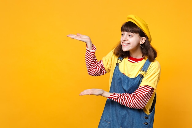 Portret van een vrolijk meisje tiener in Franse baret en denim zomerjurk wijzende handen, opzij kijken geïsoleerd op gele muur achtergrond. Mensen oprechte emoties, lifestyle concept. Bespotten kopie ruimte.