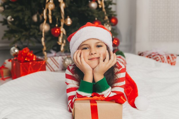 Portret van een vrolijk meisje van 8 jaar oud liggend op de vloer in de buurt van dozen met geschenken en een kerstboom thuis.