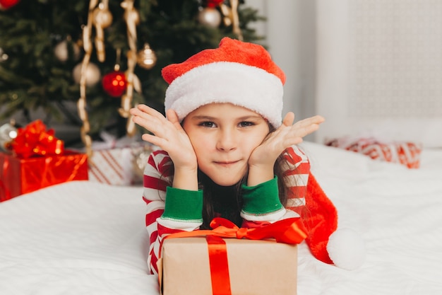 Portret van een vrolijk meisje van 8 jaar oud liggend op de vloer in de buurt van dozen met geschenken en een kerstboom thuis.