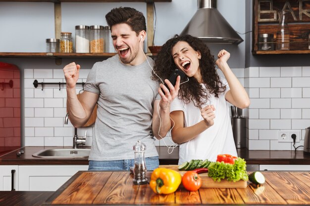 Portret van een vrolijk paar man en vrouw die samen naar muziek luisteren terwijl ze thuis salat in de keuken koken