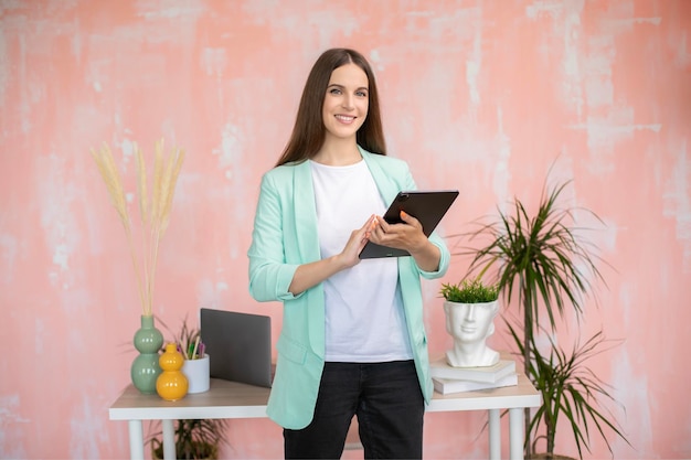Portret van een vrolijk, stijlvol meisje in een slimme, casual outfit die aan het bureau staat met een tablet in handen
