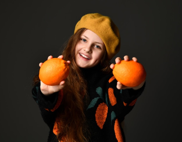 Portret van een vrolijk tienermeisje met sinaasappels die op een zwarte achtergrond staan Kind met mooi lang haar met gele baret en trui met oranje print