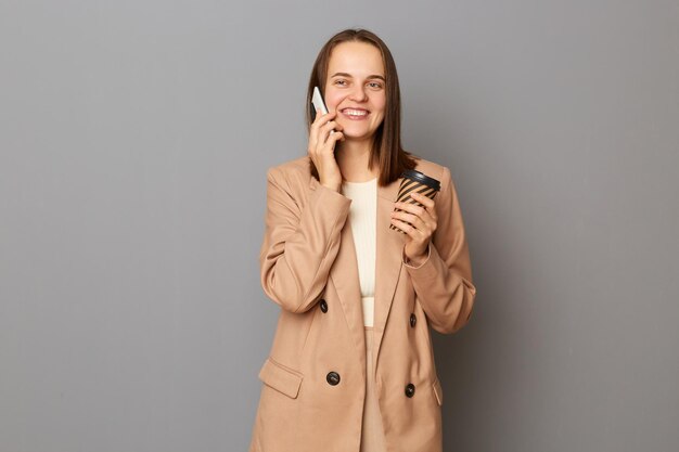 Portret van een vrolijke, aantrekkelijke, gelukkige vrouw, gekleed in een beige jas, geïsoleerd over een grijze achtergrond, afhaalkoffie drinkend en pratend op een mobiele telefoon die positieve emoties uitdrukt