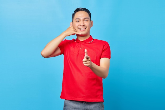 Portret van een vrolijke Aziatische jongeman die een gebaar van een telefoonteken doet en met de vinger wijst naar de camera die op een blauwe achtergrond wordt geïsoleerd