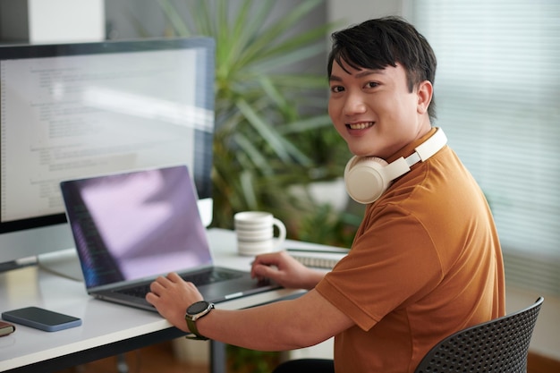 Portret van een vrolijke Aziatische softwareontwikkelaar die op de computer achter zijn bureau werkt en zich weer naar de camera wendt