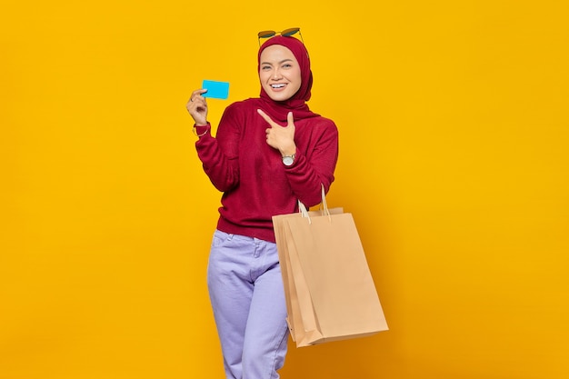 Portret van een vrolijke Aziatische vrouw die een boodschappentas vasthoudt en een creditcard toont op een gele achtergrond