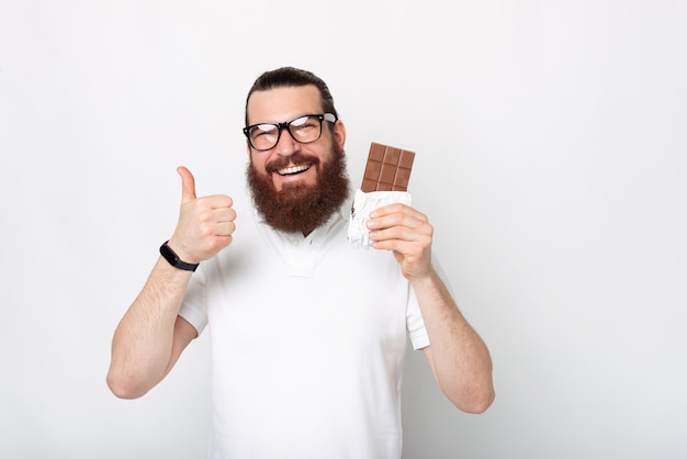 Portret van een vrolijke bebaarde man in een wit t-shirt met duim omhoog en een reep chocolade