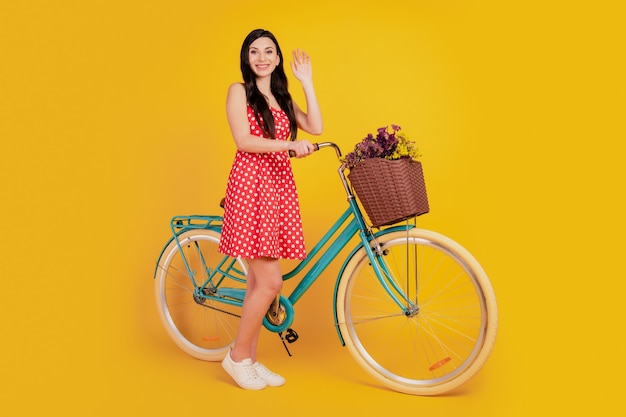 Portret van een vrolijke dame die met de fiets golft, draagt een gestippelde rode mini-jurk op een gele achtergrond
