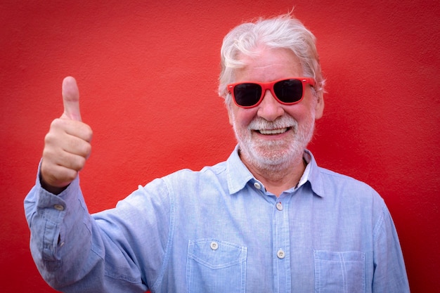 Portret van een vrolijke en lachende senior witharige man die naar de camera kijkt met een duim omhoog die tegen een rode achtergrond staat.