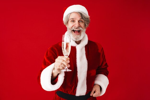 Portret van een vrolijke grijsharige oude man van de kerstman die lacht en champagneglas vasthoudt dat op rood in de studio wordt geïsoleerd