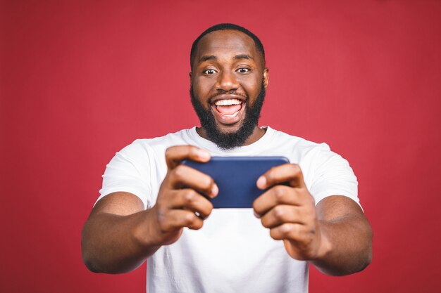 Portret van een vrolijke jonge afrikaanse man gekleed in casual spelletjes spelen op mobiele telefoon geïsoleerd op rode achtergrond.