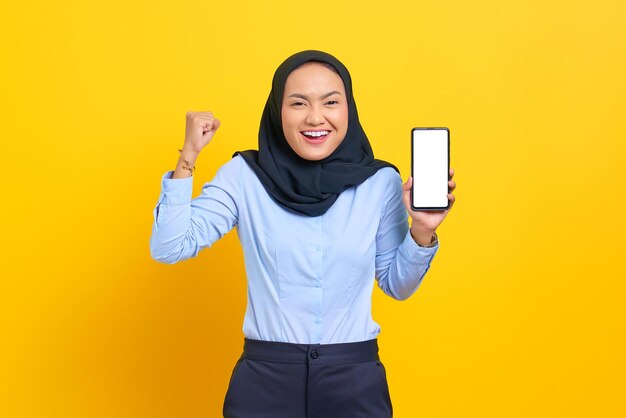 Portret van een vrolijke jonge Aziatische vrouw die een mobiele telefoon met een leeg scherm toont en een succesgebaar maakt dat op een gele achtergrond wordt geïsoleerd