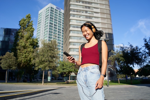 Portret van een vrolijke jonge aziatische vrouw die op straat in de stad naar muziek luistert of video's bekijkt op haar mobiele telefoon. kijkend naar de camera. hoge kwaliteit foto