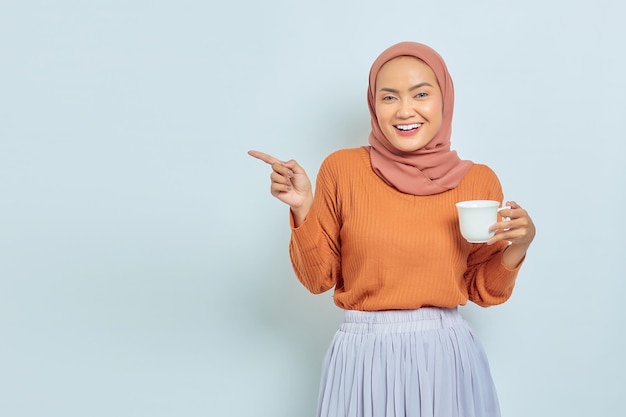 Portret van een vrolijke jonge Aziatische vrouw in een bruine trui die een beker vasthoudt en wijst om de ruimte te kopiëren met een vinger geïsoleerd op een witte achtergrond