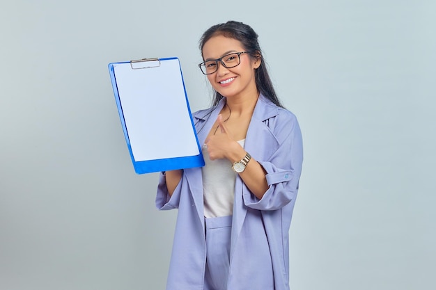 Portret van een vrolijke jonge Aziatische zakenvrouw die een leeg klembord en een duimgebaar toont dat op een paarse achtergrond wordt geïsoleerd