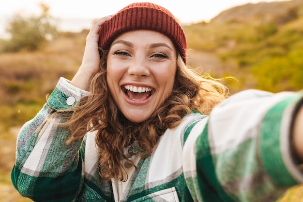 Portret van een vrolijke jonge blanke vrouw met een hoed en een geruit hemd die een selfie neemt terwijl ze buiten loopt