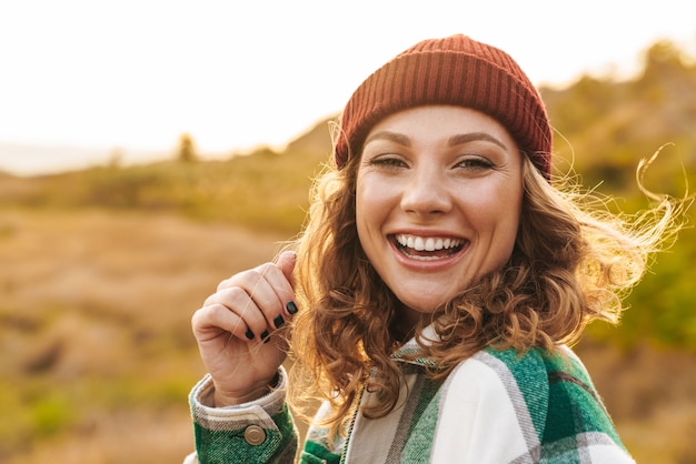 Portret van een vrolijke jonge blanke vrouw met een hoed en een geruit hemd die lacht terwijl ze buiten loopt