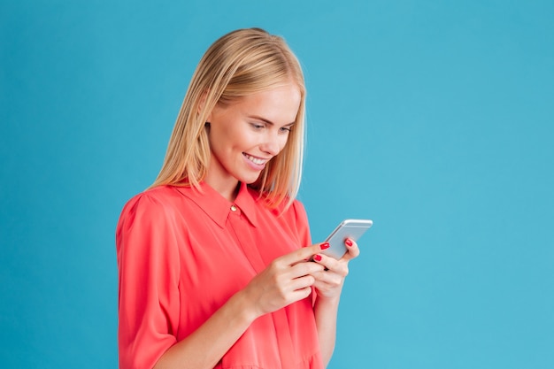 Portret van een vrolijke jonge blonde vrouw die mobiele telefoon over blauwe muur houdt