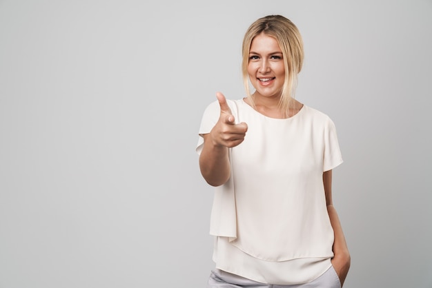 Portret van een vrolijke jonge geweldige blonde vrouw die zich geïsoleerd over een grijze muur poseert, gekleed in een basic wit t-shirt dat naar jou wijst