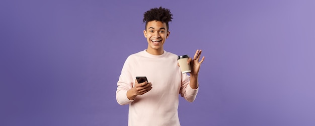 Portret van een vrolijke jonge mannelijke student met een kopje koffie en een mobiele telefoon die met de hand zwaait om hallo te zeggen