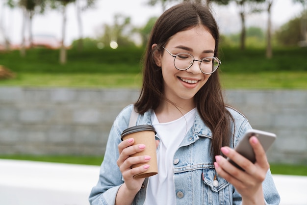 Portret van een vrolijke jonge mooie vrouwelijke student die buiten in het groene natuurpark loopt en koffie drinkt met behulp van chatten op de mobiele telefoon