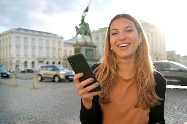 Portret van een vrolijke jonge vrouw die lacht naar de camera met smartphone in Brussel, België Sunset city lifestyle-technologie