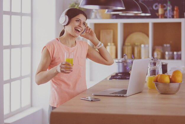 Portret van een vrolijke jonge vrouw die naar muziek luistert met een koptelefoon en een laptop gebruikt terwijl ze in de keuken staat